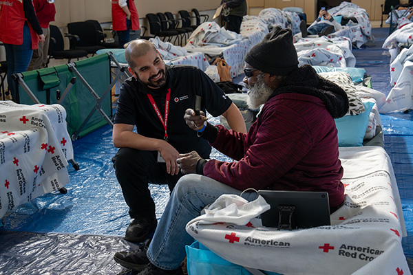 A Red Cross worker and an individual are interacting at a shelter with beds and blankets marked with the American Red Cross logo. The worker is kneeling and smiling while talking to the seated person, who is holding a cellphone.