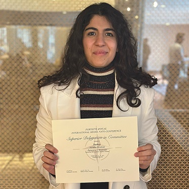 Individual holding a certificate from the International Model United Nations Conference on the Syrian Refugee Committee, smiling in an indoor setting.