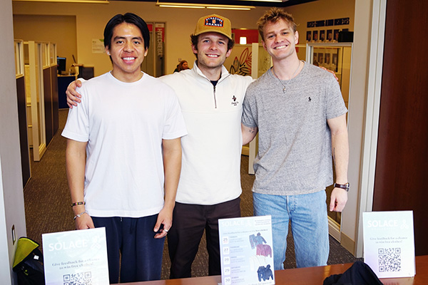 Three individuals standing side by side in an office environment, smiling at the camera, with posters titled