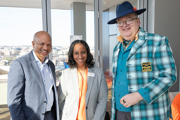 Three individuals smiling indoors at an event with a cityscape visible through the windows behind them. The person on the right is wearing a plaid suit and a blue hat with a Maxwell badge.