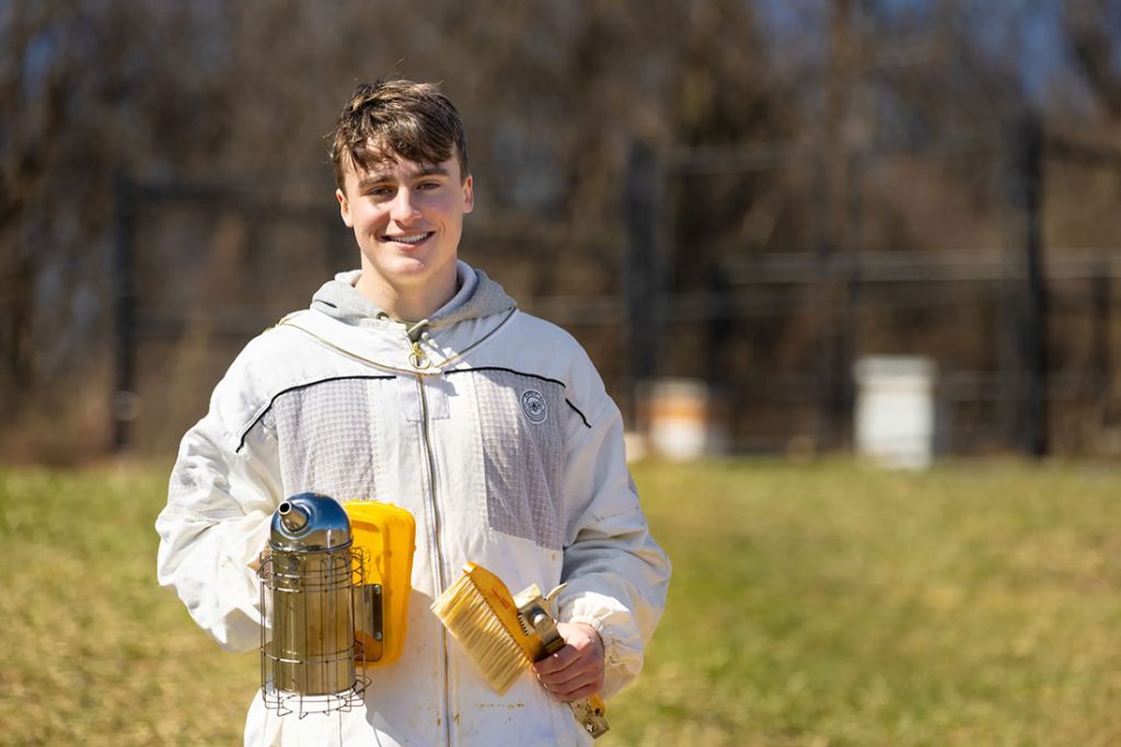 male student holding bee keeping equipment and smiling
