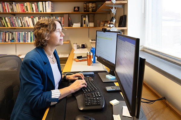 A professional is working at a desk in an office, using a computer with dual monitors displaying graphs and text. The office is filled with books and personal items, conveying a productive environment.