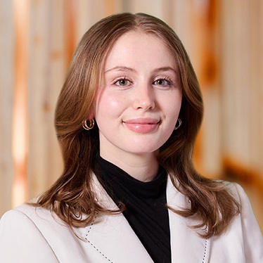 Portrait of a smiling person with shoulder-length hair, wearing a white jacket and black turtleneck, standing in front of a blurred wooden background.