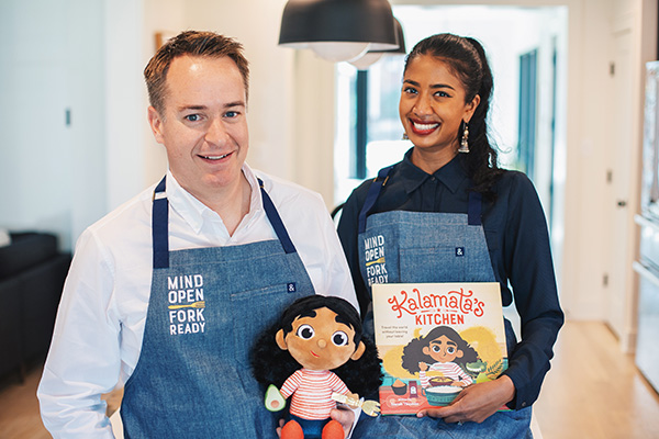 Two individuals wearing aprons stand in a kitchen, holding a book titled