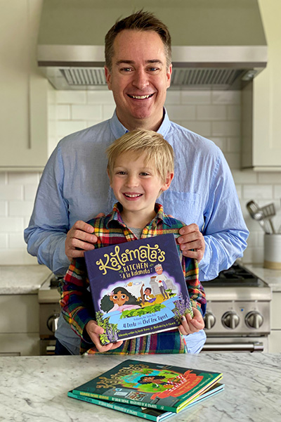 Two individuals standing in a kitchen, one adult and one child, holding a book titled