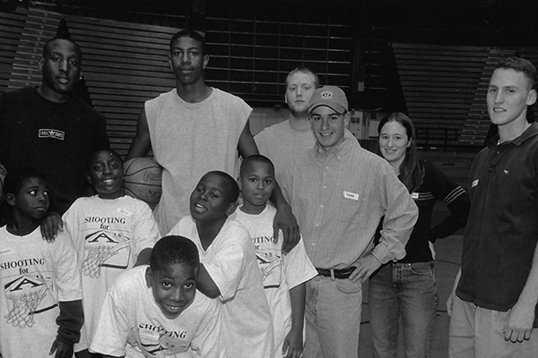 Group of diverse individuals, including children and adults, posing in a basketball gym. Some are wearing T-shirts with