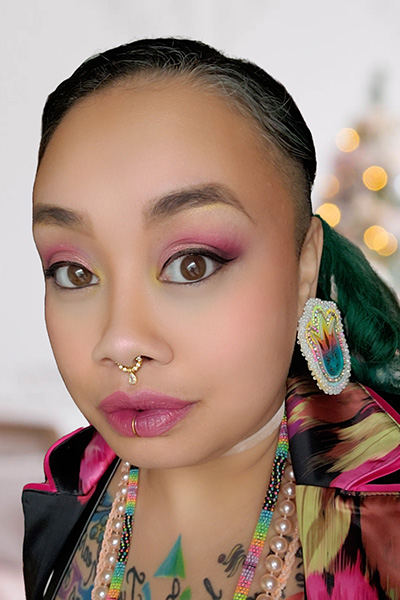 Close-up portrait of a person with intricate makeup and a nose ring, wearing colorful attire and an earring. The background is softly blurred with faint lights.
