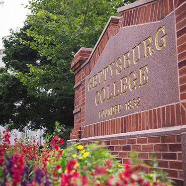 Brick sign displaying "Gettysburg College, Founded 1832" with colorful flowers in the foreground and a statue near a building in the background.