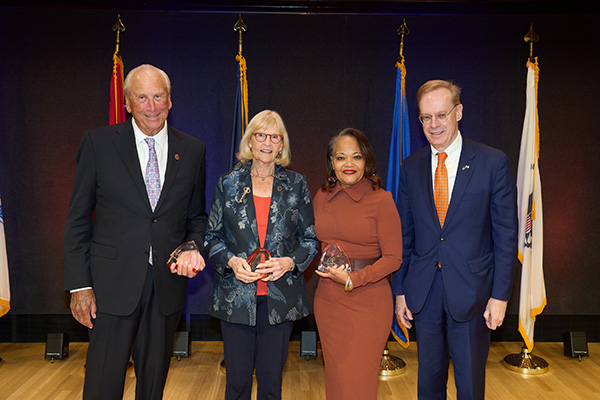 Four individuals standing side-by-side at a formal award ceremony, holding awards, with several flags in the background.