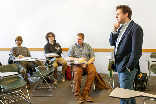 A classroom scene with four individuals focused on a speaker standing to the right, who appears thoughtful.