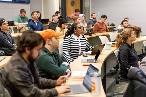 A diverse group of students sits in a university lecture hall, attentively listening to a presentation. Some are taking notes on laptops and tablets.