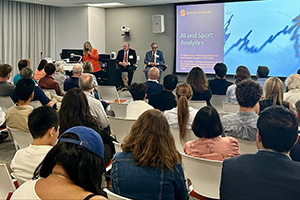 Attendees at a business seminar hosted by Syracuse University, focusing on Asset Sport Analytics, with presenters speaking at the front next to a slideshow screen.