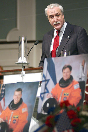Former NASA Administrator Sean O'Keefe speaking at a podium with photographs of astronauts in space suits displayed in the foreground.