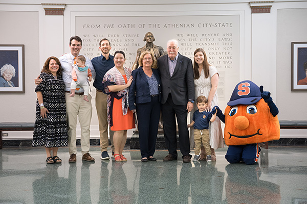 A group of eight people posing for a photo in a hallway with a text from the Athenian City-State on the wall above them. The group includes adults, children, and Otto the Orange, the Syracuse University mascot.