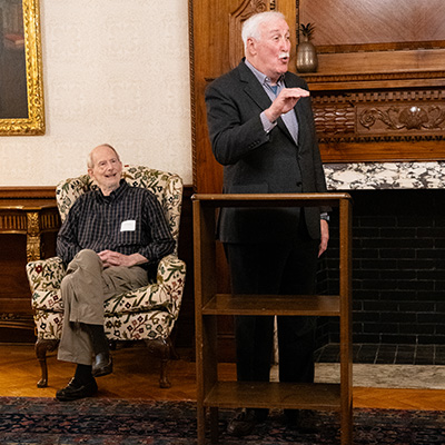 Two individuals in a formal setting, one seated and one standing and speaking, with an ornate fireplace in the background.