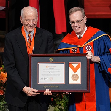 Two individuals smiling at a graduation ceremony, with one presenting an award to the other, who is wearing academic regalia representing Syracuse University.