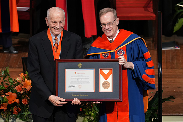 Two individuals smiling at a graduation ceremony, with one presenting an award to the other, who is wearing academic regalia representing Syracuse University.