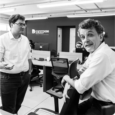 Two individuals in an office setting at DEEP, engaged in a conversation with computers and colleagues in the background.