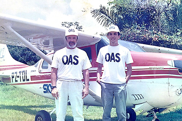 Two individuals wearing 'OAS' caps and t-shirts stand in front of a small red and white airplane, posing on a grassy field.