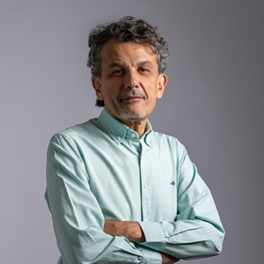 Portrait of an individual standing with arms crossed, wearing a light green shirt with a small Lacoste logo, against a gray backdrop.