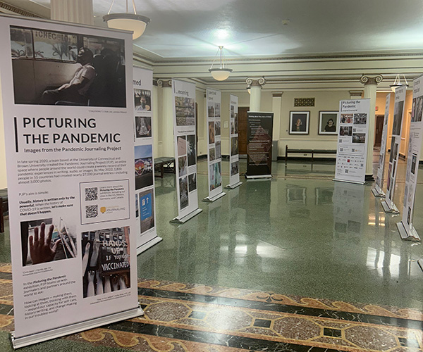Exhibition titled 'Picturing the Pandemic' with informational panels displayed in a hallway, featuring photographs and text about the global impact of the pandemic.