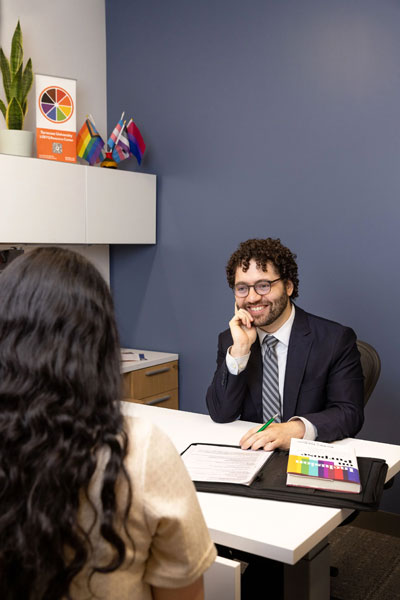 A student in a suit sits across from another student in an office.