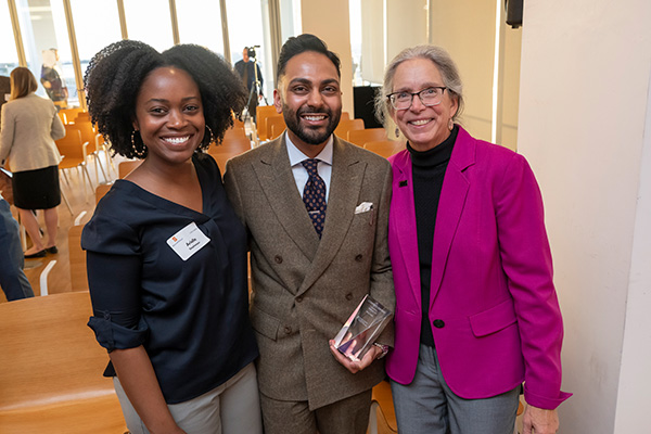 hree professionals smiling at a networking event, one holding an award. The setting includes a bright room with chairs and tables in the background.