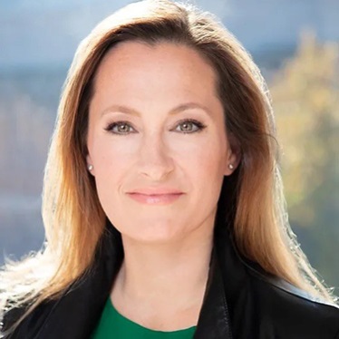 A professional headshot of an individual with shoulder-length light brown hair, wearing a black jacket and a green top, with a blurred cityscape background.