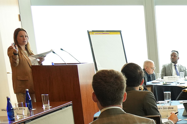 A person presents at a podium to an audience of professionals seated at tables during a conference. A whiteboard and a large window with a view of the skyline are visible in the background.