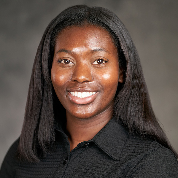 A portrait of a smiling individual with long black hair, wearing a black shirt. The background is plain and gray.