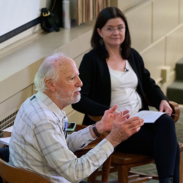 Robert Shetterly, seated and gesturing during a discussion, with an attentive audience member next to him.