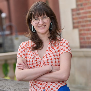 Person in a red and white floral shirt and glasses, smiling with arms crossed, standing in front of a brick building.