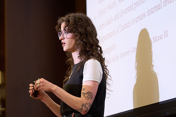 A person presenting at a conference, standing at a podium with a microphone, in front of a screen displaying text.