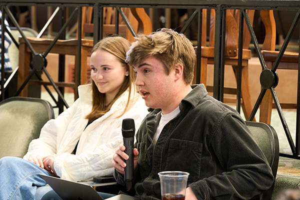 Two people sitting at a discussion panel, one holding a microphone and speaking, with attentive expressions in an indoor setting.