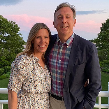 Two people smiling for a photo on a balcony with a scenic sunset in the background.