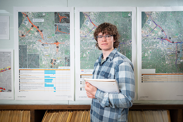 Person holding a clipboard stands in front of wall-mounted maps and charts in a workspace.