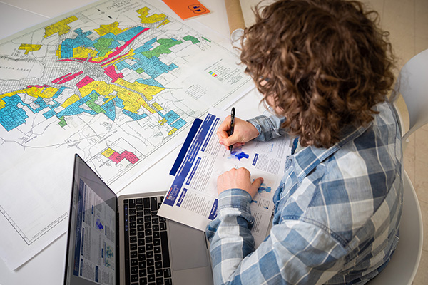 Person working at a desk with a laptop, writing in a notebook, with a large colorful map spread out on the table.