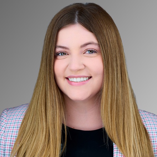 Professional headshot of a smiling individual with long blond hair, wearing a tweed jacket against a neutral background.