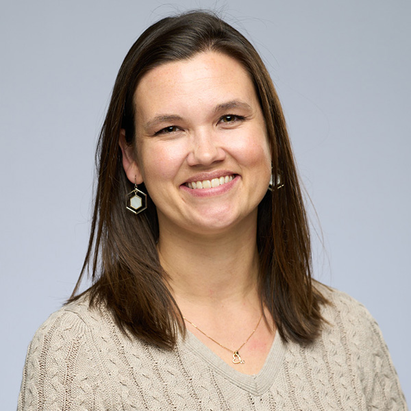 Portrait of a smiling person wearing a beige sweater and hexagonal earrings, set against a plain gray background.