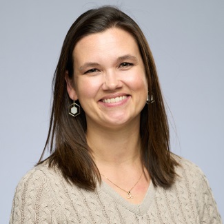Portrait of a smiling person wearing a beige sweater and hexagonal earrings, set against a plain gray background.