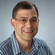 Professional headshot of a smiling individual wearing glasses and a striped shirt.
