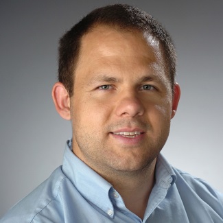 Professional headshot of a person with a short haircut, wearing a light blue shirt, smiling against a gray background.