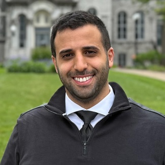 A smiling individual standing in front of a historic stone building.