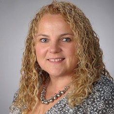 Portrait of an individual with curly blonde hair, wearing a patterned top and a silver necklace, smiling at the camera.