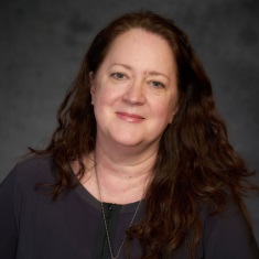 Portrait of a smiling individual with long, curly hair, wearing a dark top and a necklace, against a gray background.